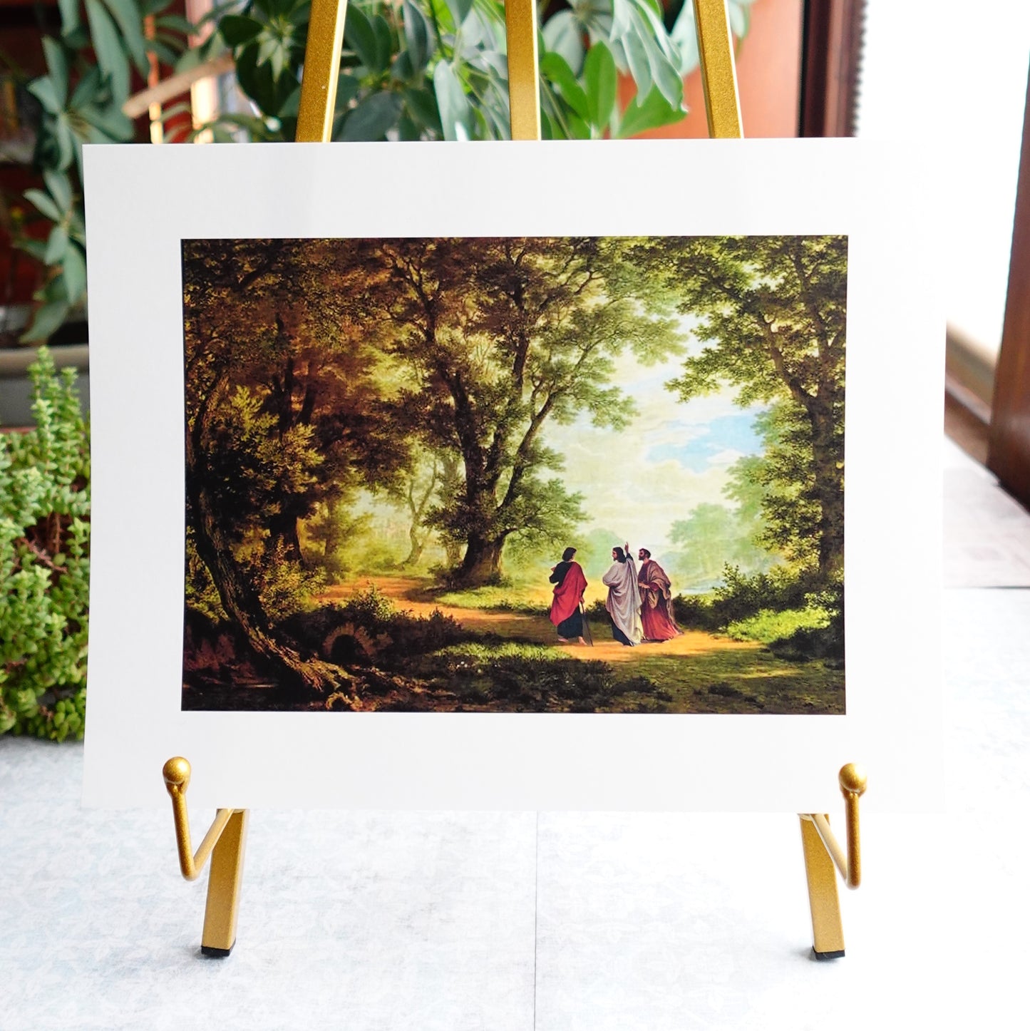 The image is a framed print of a painting depicting a group of people walking through a lush, forested area.