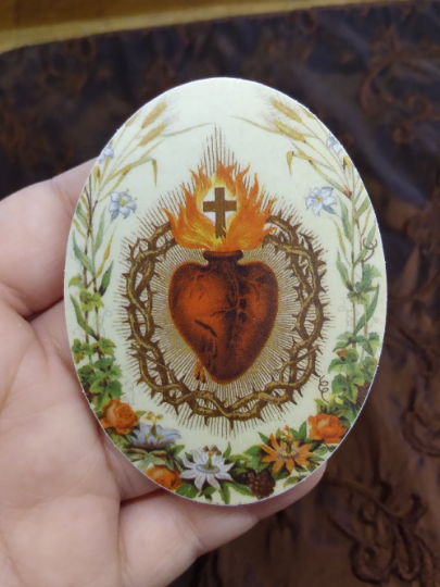 A hand holding a small oval-shaped object with a religious symbol on it, surrounded by flowers and a wreath.