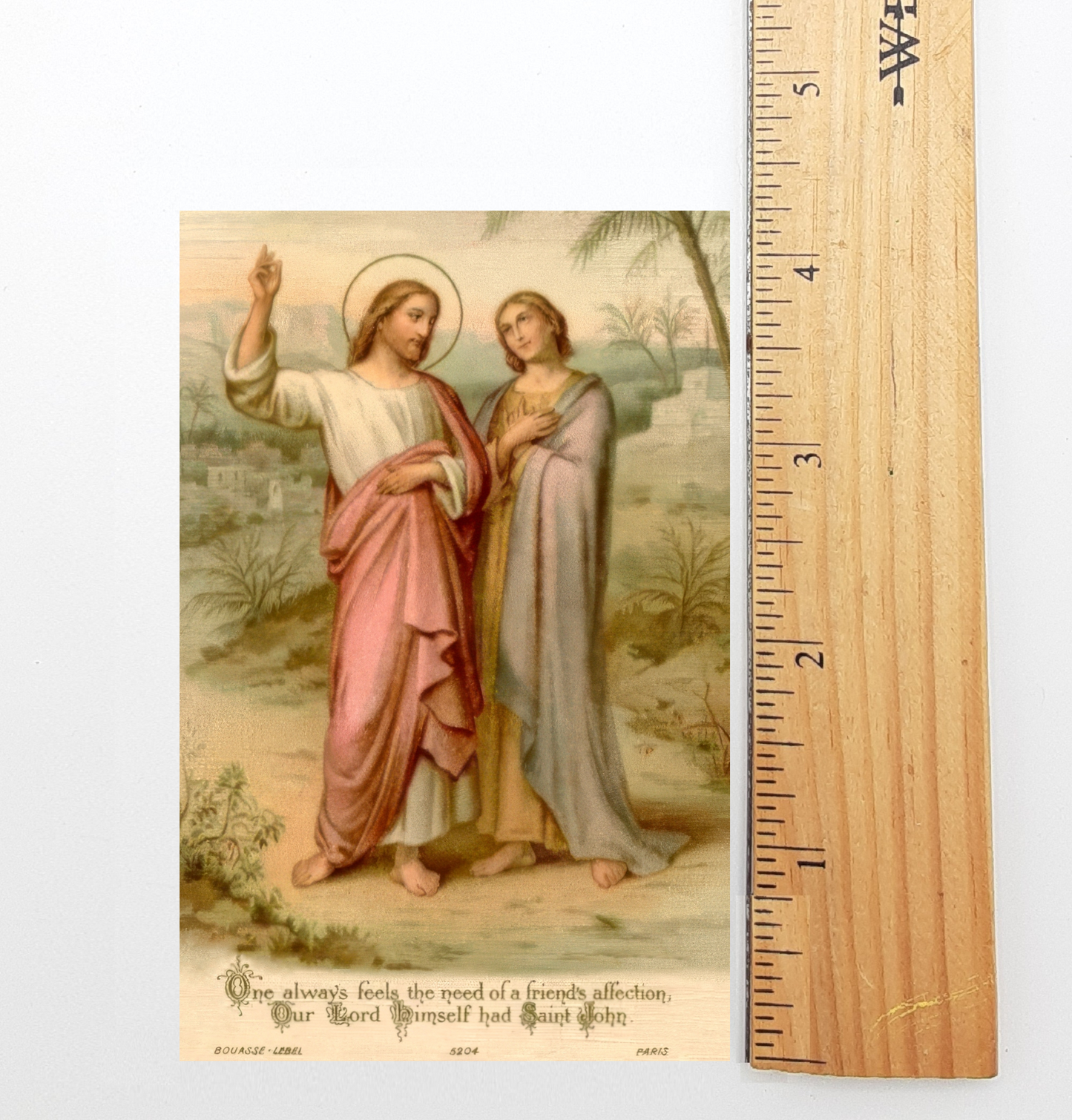 The image is a religious painting depicting two figures, likely Jesus and Mary, standing in a natural setting. The painting is framed by a wooden ruler, which is marked with measurements in inches. The text below the painting reads "One always feels the need of a friendly affection. Our Lord Himself had said, 'Be kind to one another.' BOUQUET-LEBEL, PARIS, 624."