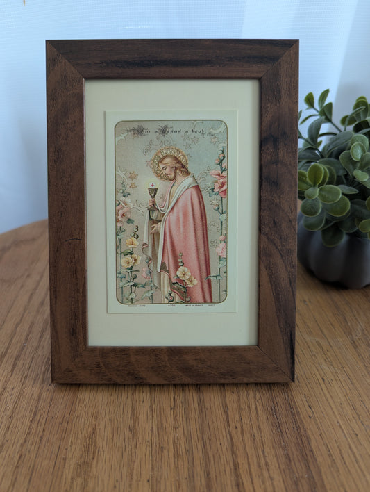 a framed religious illustration of a person in a pink robe, holding a chalice, surrounded by flowers.