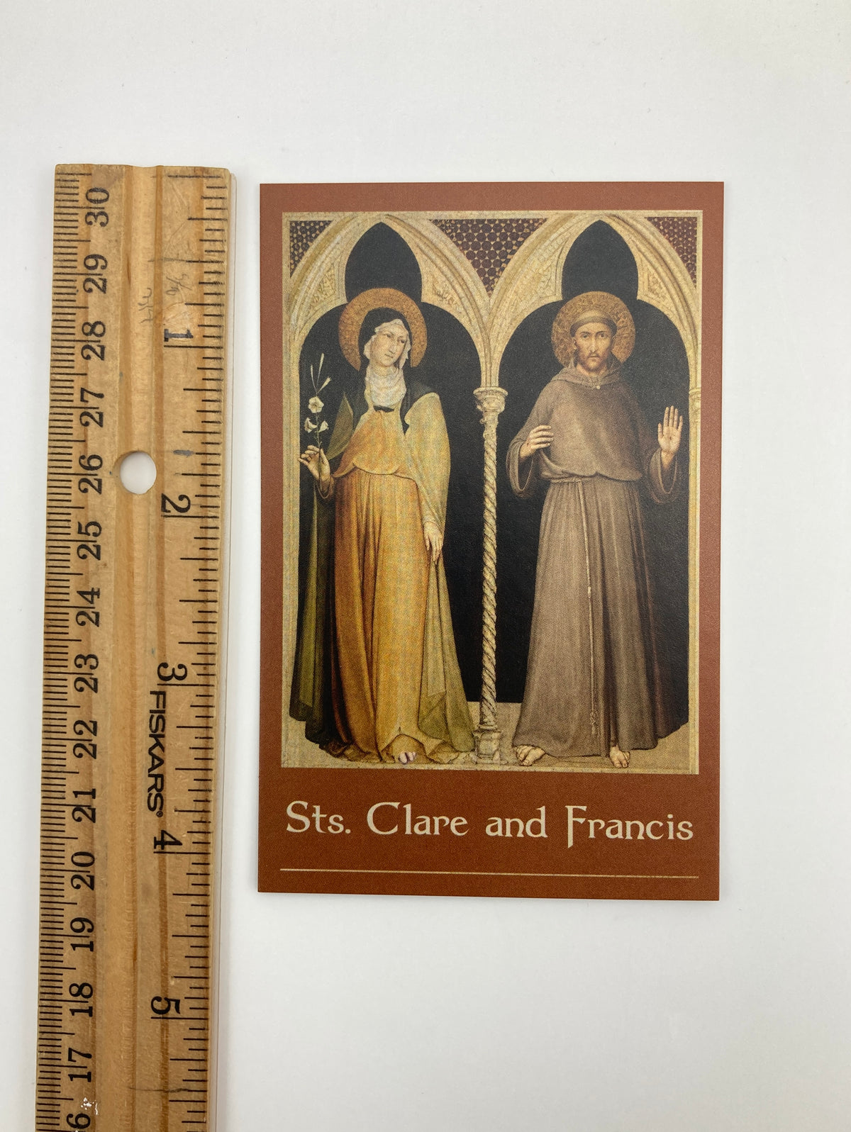 A brown-framed card with an illustration of two religious figures, likely Sts. Clare and Francis, standing in front of a cross. The card is placed next to a ruler for scale.