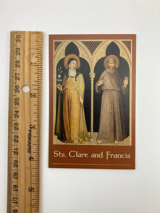 A brown-framed card with an illustration of two religious figures, likely Sts. Clare and Francis, standing in front of a cross. The card is placed next to a ruler for scale.