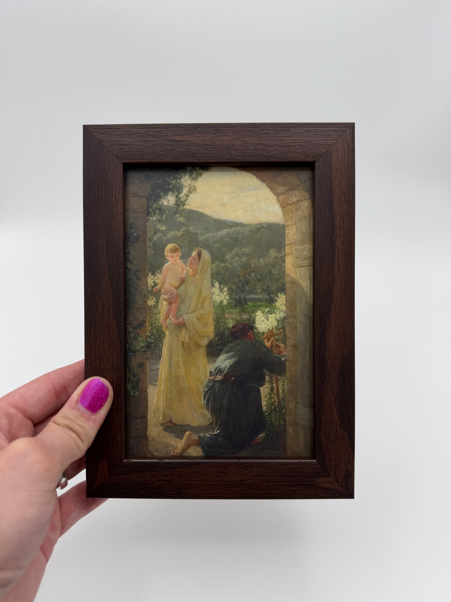 A hand holding a small framed painting of a woman in a yellow dress, with a child in her arms, against a natural background.