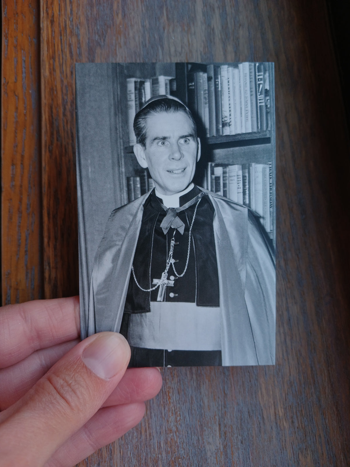 A hand is holding a black and white photograph of a smiling man wearing a robe and standing in front of a bookshelf.