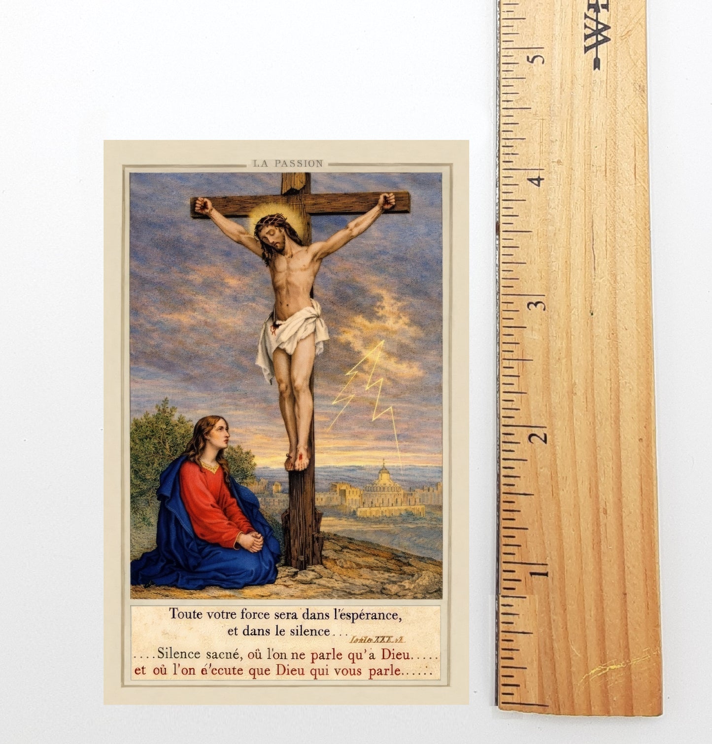 The image is a religious painting depicting the crucifixion of Jesus Christ, with a woman kneeling beside him. The painting is framed by a wooden ruler, which is used to measure the size of the artwork.