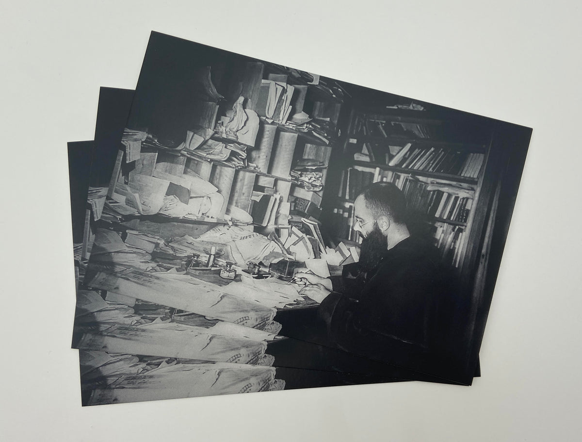 A black and white photograph of a man sitting at a desk surrounded by books and papers, with a cluttered and disorganized appearance.