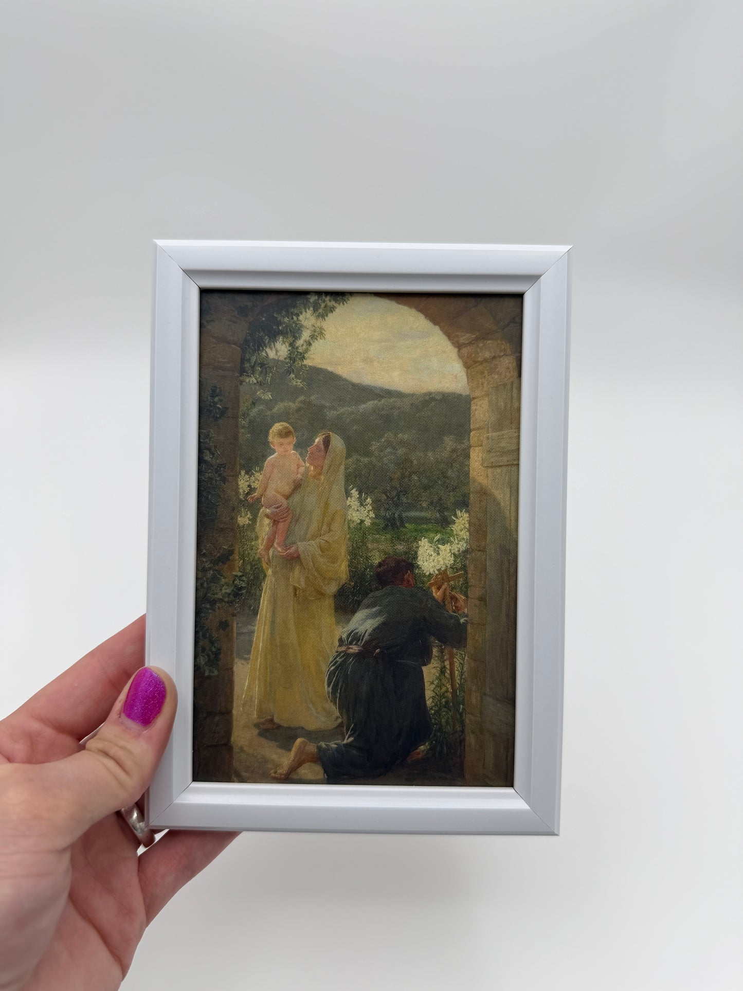 A hand holding a small framed painting of a woman in a yellow dress, with a man in a black robe standing next to her.
